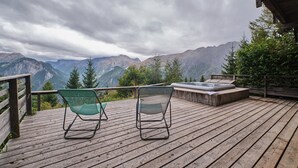 Une terrasse en bois avec deux chaises vertes et un jacuzzi, le tout offrant une vue sur un paysage montagneux.
