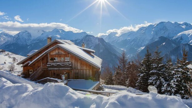 Eine Holzchalet mit Balkon, umgeben von schneebedeckten Bergen und blauem Himmel.