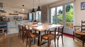 Een moderne keuken met een grote eettafel, houten stoelen, hanglampen en uitzicht door grote ramen.