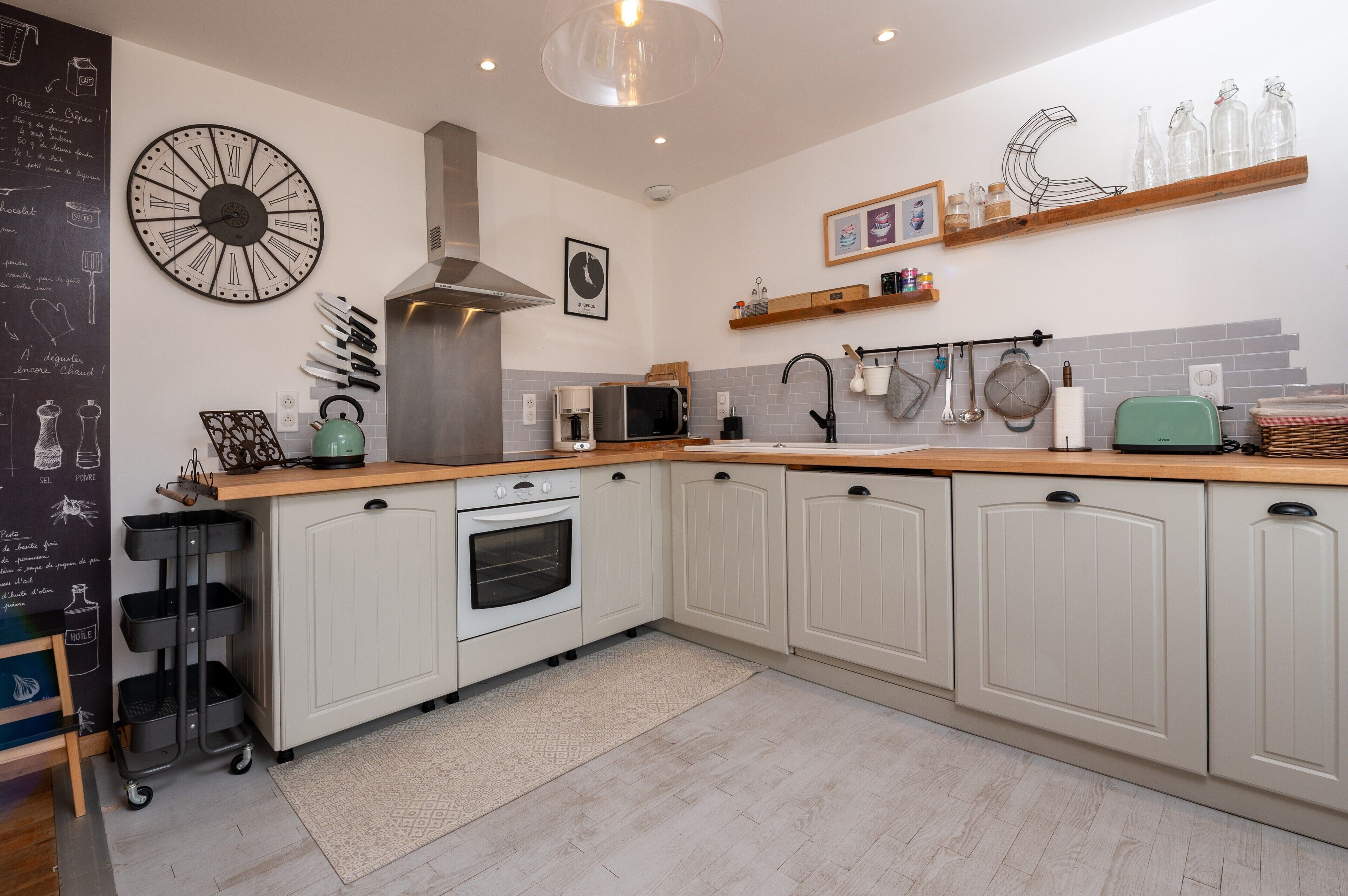A kitchen with white cabinets, a stainless steel range hood, a microwave, and a kettle. There are shelves with jars and a chalkboard with writing.
