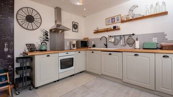 A kitchen with white cabinets, a stainless steel range hood, a microwave, and a kettle. There are shelves with jars and a chalkboard with writing.