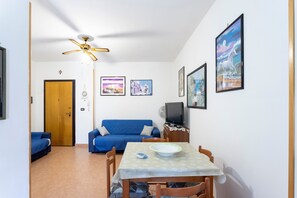Une petite pièce comprenant une table à manger, un canapé bleu, un ventilateur de plafond et un téléviseur fixé au mur.