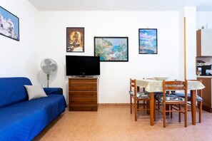 Un salon avec un canapé bleu, un téléviseur, un ventilateur, une table à manger avec des chaises et des œuvres d’art accrochées aux murs.