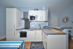 Une cuisine compacte avec des meubles blancs, un four à micro-ondes encastré, une cuisinière et un petit îlot central doté d’une planche à découper.