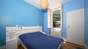 A bedroom with a bed, white nightstands, a window with a view of greenery, and a white door.