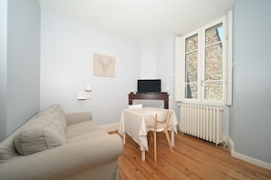 A room with a sofa, a table with chairs, a TV on a stand, a radiator, and a window with a stone wall view.