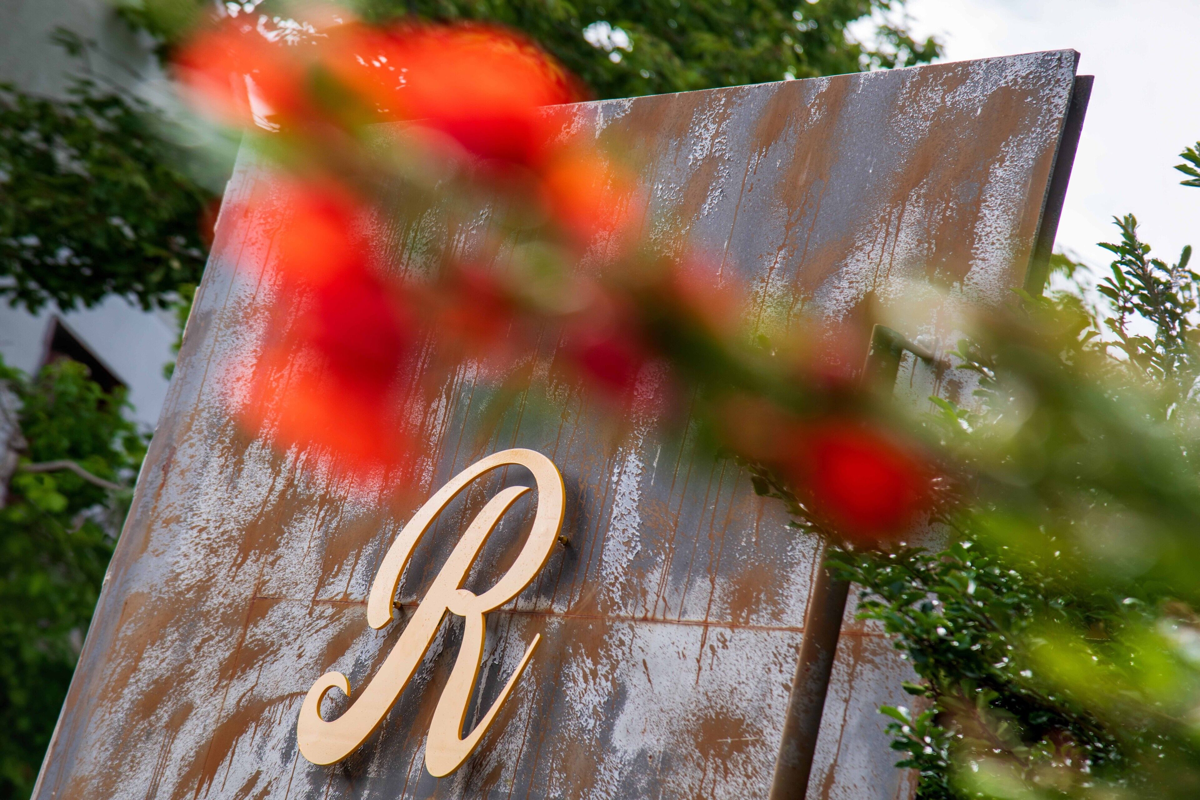 Een metalen bord met een grote, versierde letter 'R' en een gedeeltelijk zicht op een gebouw op de achtergrond.