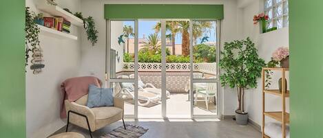 Una habitación con sofá, una silla, una alfombra pequeña, una planta en maceta y vistas a un patio exterior con piscina y palmeras.