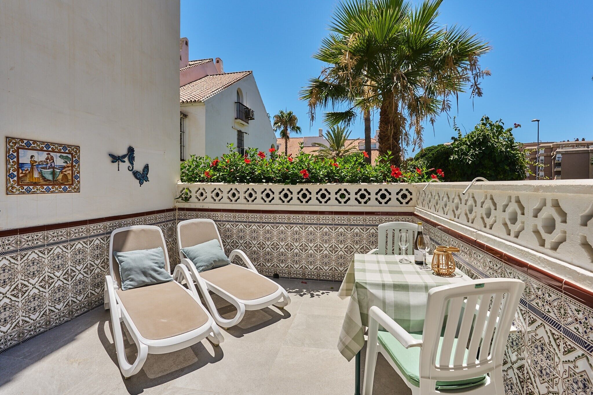Un patio con dos sillones de descanso y una mesa, rodeado de azulejos decorativos y vistas a un edificio y palmeras.