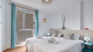 A bedroom with two beds, a window with curtains, a bedside table with a lamp, and a wall-mounted decorative item.