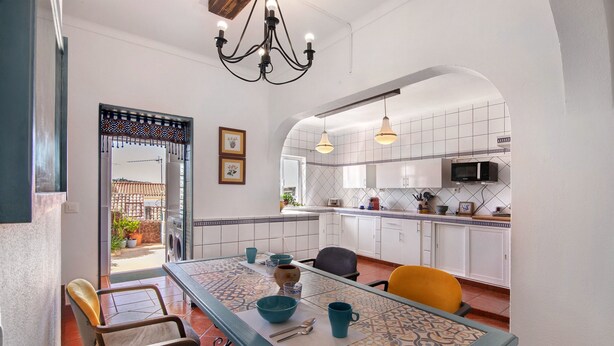 Een eethoek met een tafel voor vier personen, een keuken met witte kasten en een betegelde spatwand, en een kroonluchter erboven.