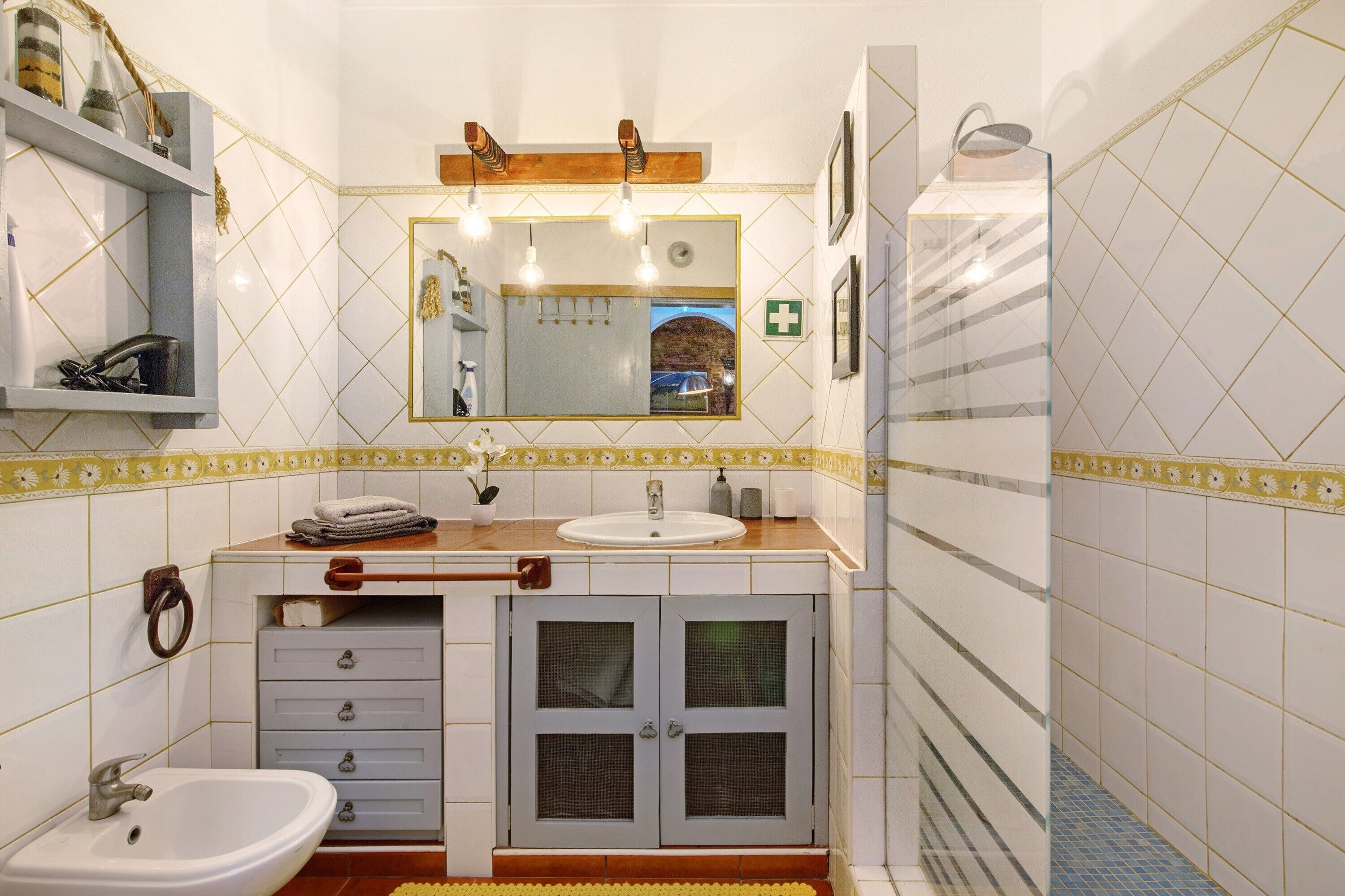 Une salle de bain équipée d’un lavabo, d’un miroir et d’une douche. On y trouve des étagères avec des articles de toilette et un porte-serviettes.