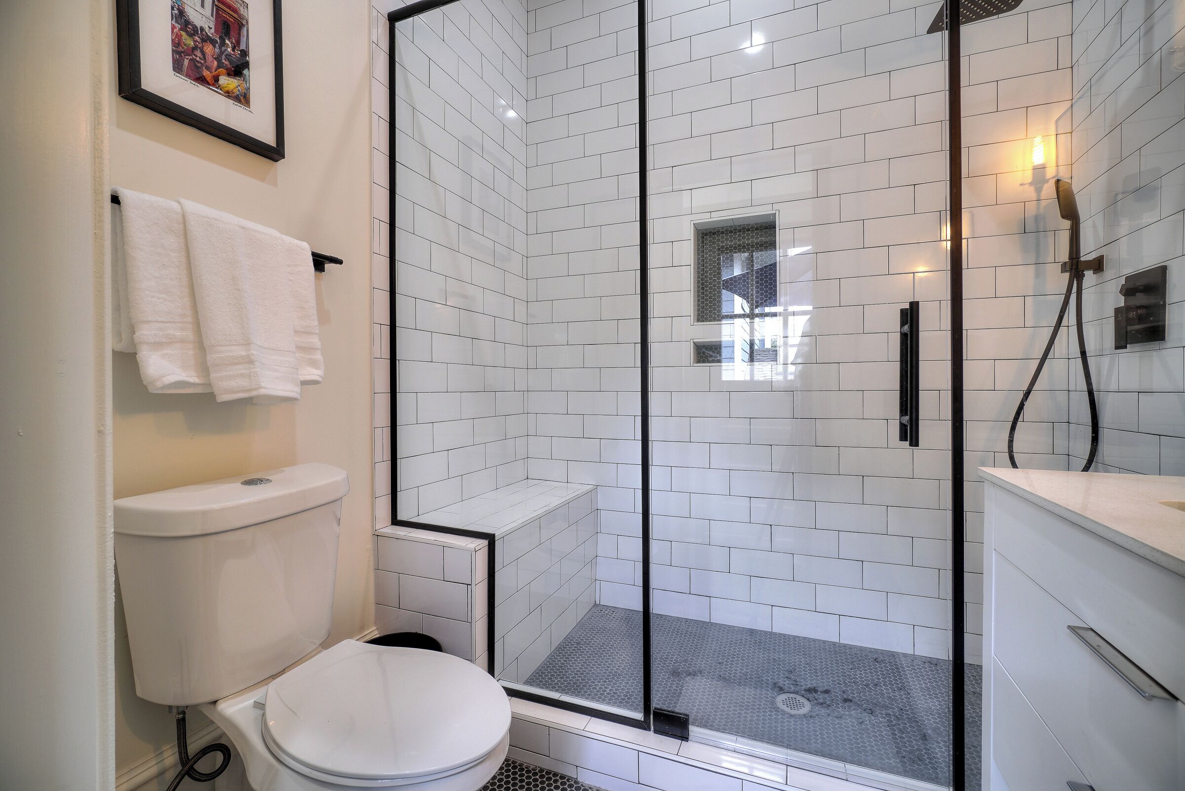 Une salle de bain moderne avec une douche à parois de verre, des murs recouverts de carreaux blancs, des toilettes et un lavabo avec un meuble-vasque.