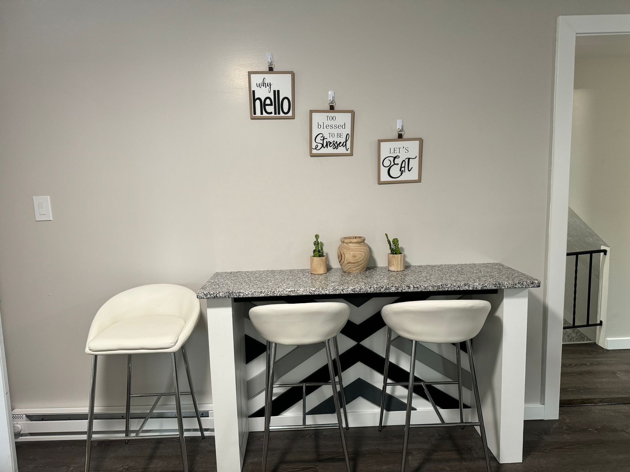Une cuisine dotée d’un comptoir en granit, de trois tabourets de bar blancs et de trois panneaux décoratifs fixés au mur, portant des phrases inspirantes.