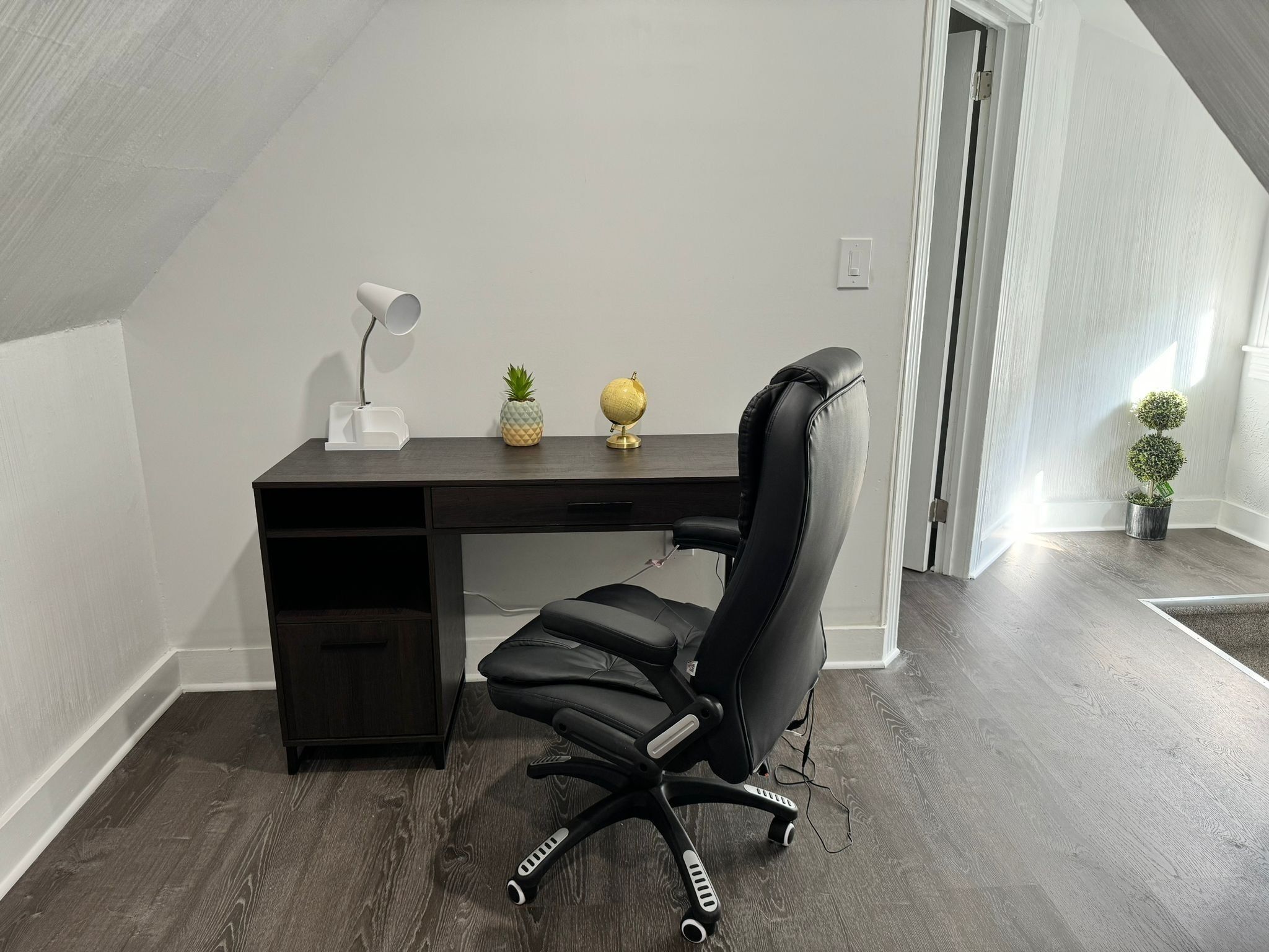 Un espace de bureau moderne avec un bureau en bois foncé, une chaise de bureau noire, une lampe de bureau blanche, un petit ananas et un globe terrestre.