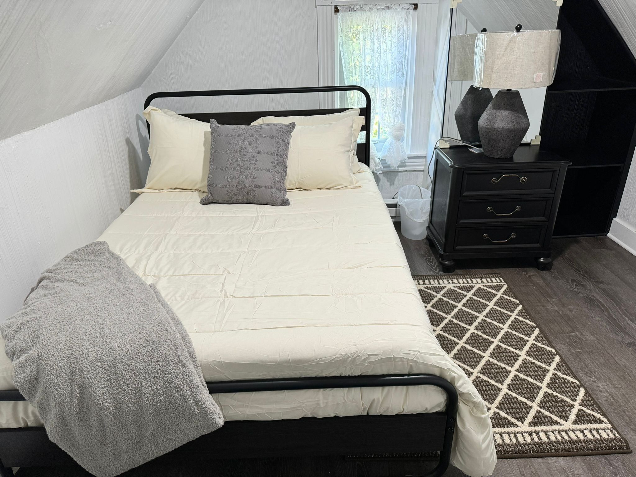 Uma cama bem arrumada com edredom branco e manta cinza, uma mesa de cabeceira com abajur e um tapete estampado sobre o assoalho de madeira.
