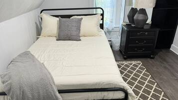 Uma cama bem arrumada com edredom branco e manta cinza, uma mesa de cabeceira com abajur e um tapete estampado sobre o assoalho de madeira.