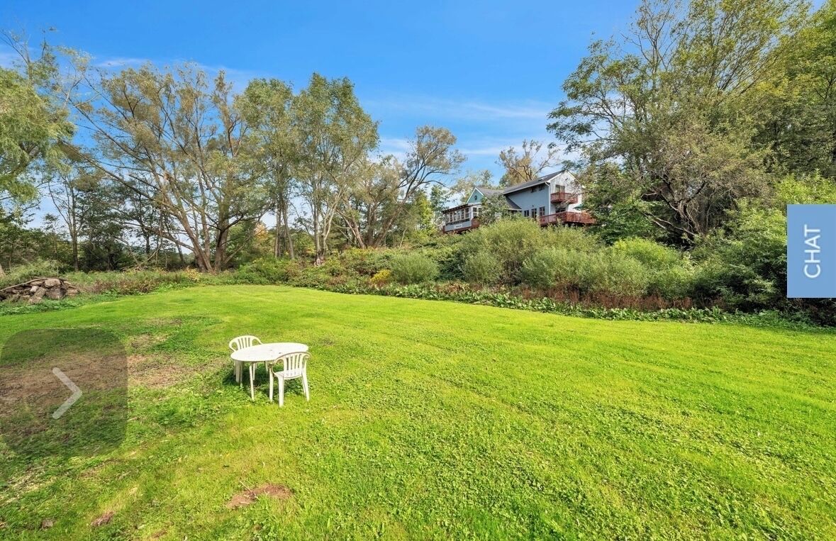 A grassy backyard with a white table and chairs, surrounded by trees and a house in the background.
