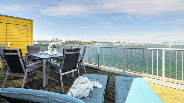 Una terraza con mobiliario exterior, una mesa y sillas, y vistas al agua y al paisaje urbano.