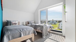 Un dormitorio con un ventanal grande, una cama con colcha gris y vistas a una zona residencial.