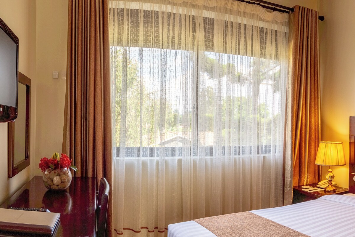 A hotel room with a bed, two bedside lamps, a nightstand, a mirror, and a window with sheer curtains.