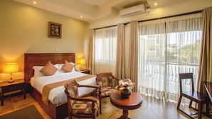 A hotel room with a large bed, two bedside lamps, a wooden headboard, a small table with a vase of flowers, a chair, and a balcony with curtains. - Nyaika Hotel Limited (Fort Portal)