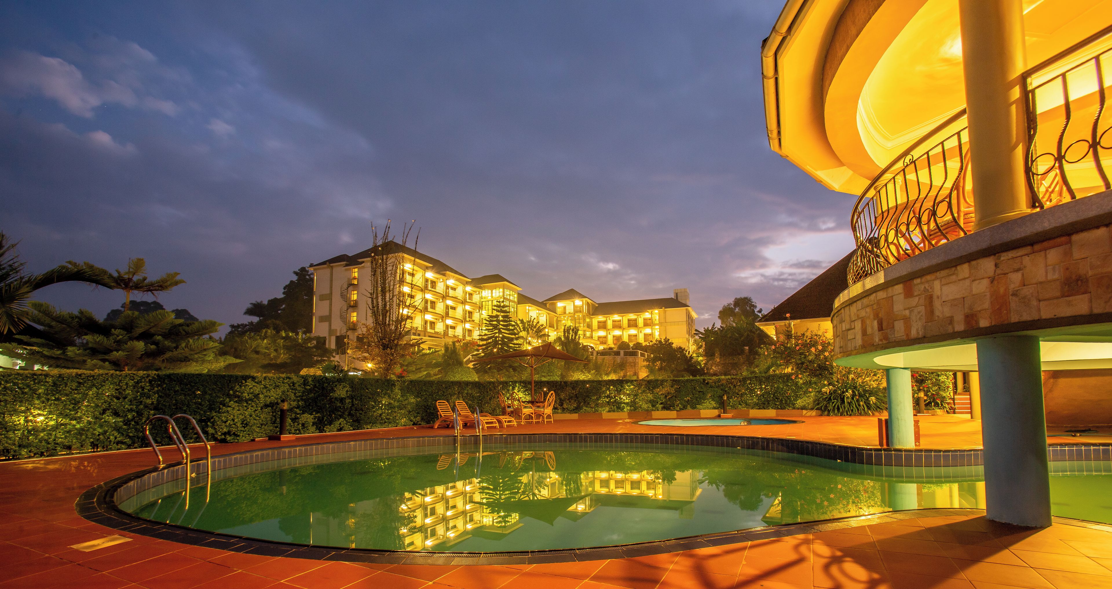 A swimming pool with a reflection of a building, surrounded by greenery and a well-lit balcony.