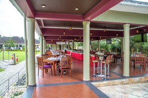 Un área de comedor al aire libre con mesas y sillas, techo cubierto y una pantalla de televisión.