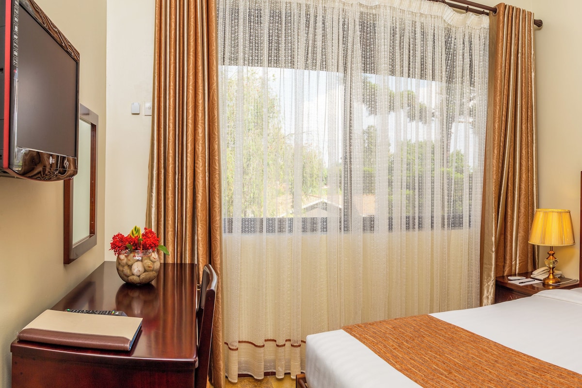 A hotel room with a large bed, wooden headboard, two bedside tables with lamps, a TV on the left, and a window with sheer curtains.