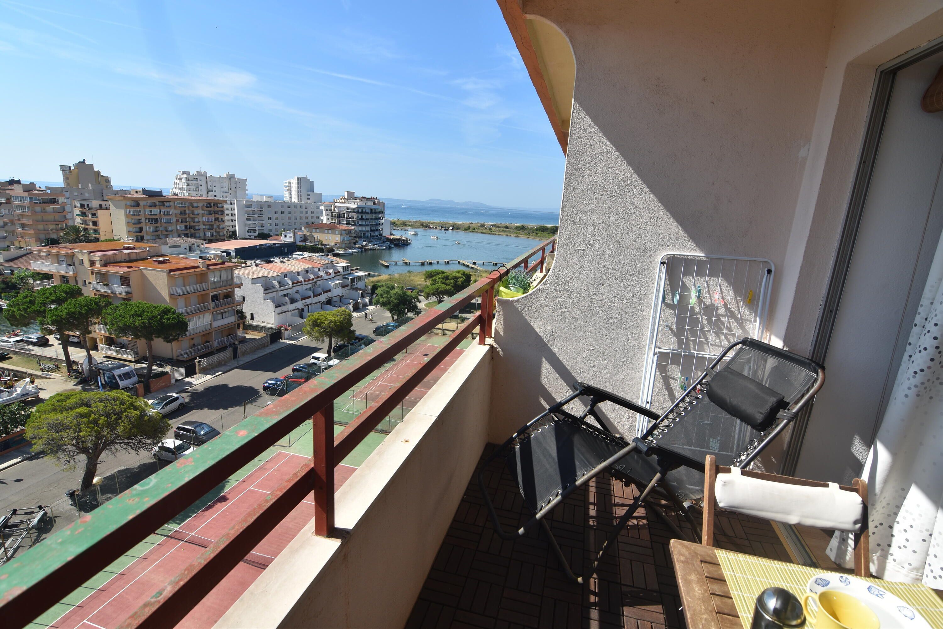 A balcony with a view of a cityscape and a body of water. There are two chairs and a table with a cup and saucer.