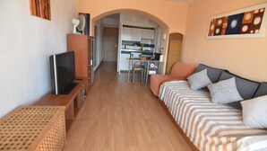 Ein kompaktes Hotelzimmer mit Fernseher, einer Couch, einem Esstisch und einer durch einen Bogen sichtbaren Küchenzeile.