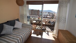 Ein Balkon mit Blick auf Gebäude und Berge, eine Couch mit gestreifter Bettwäsche und ein Hängekorbsessel.