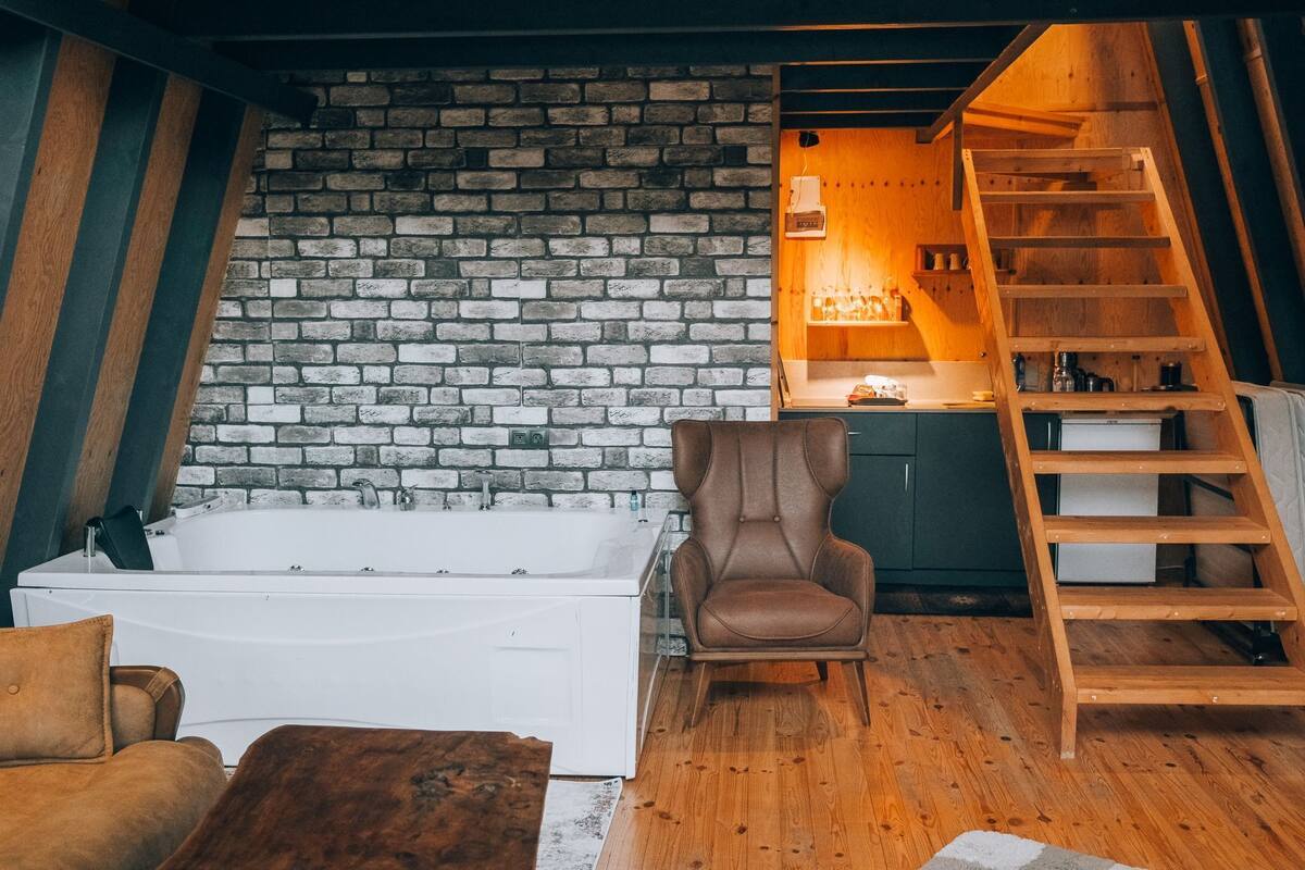 A cozy room with a brick wall, a wooden ladder, a brown leather chair, a white bathtub, and a wooden floor.