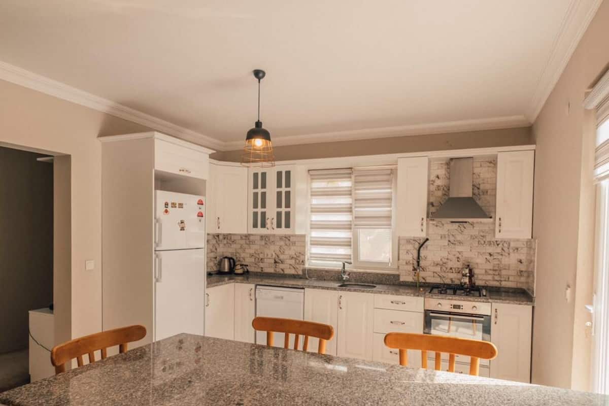 A modern kitchen with a granite island, wooden chairs, white cabinets, and a hanging light fixture.