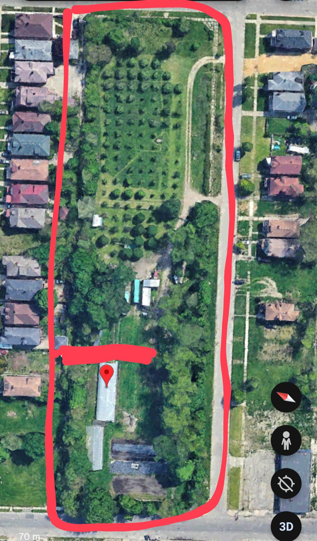 An aerial view of a residential area with a large green space, a marked location, and a road running parallel to the buildings.