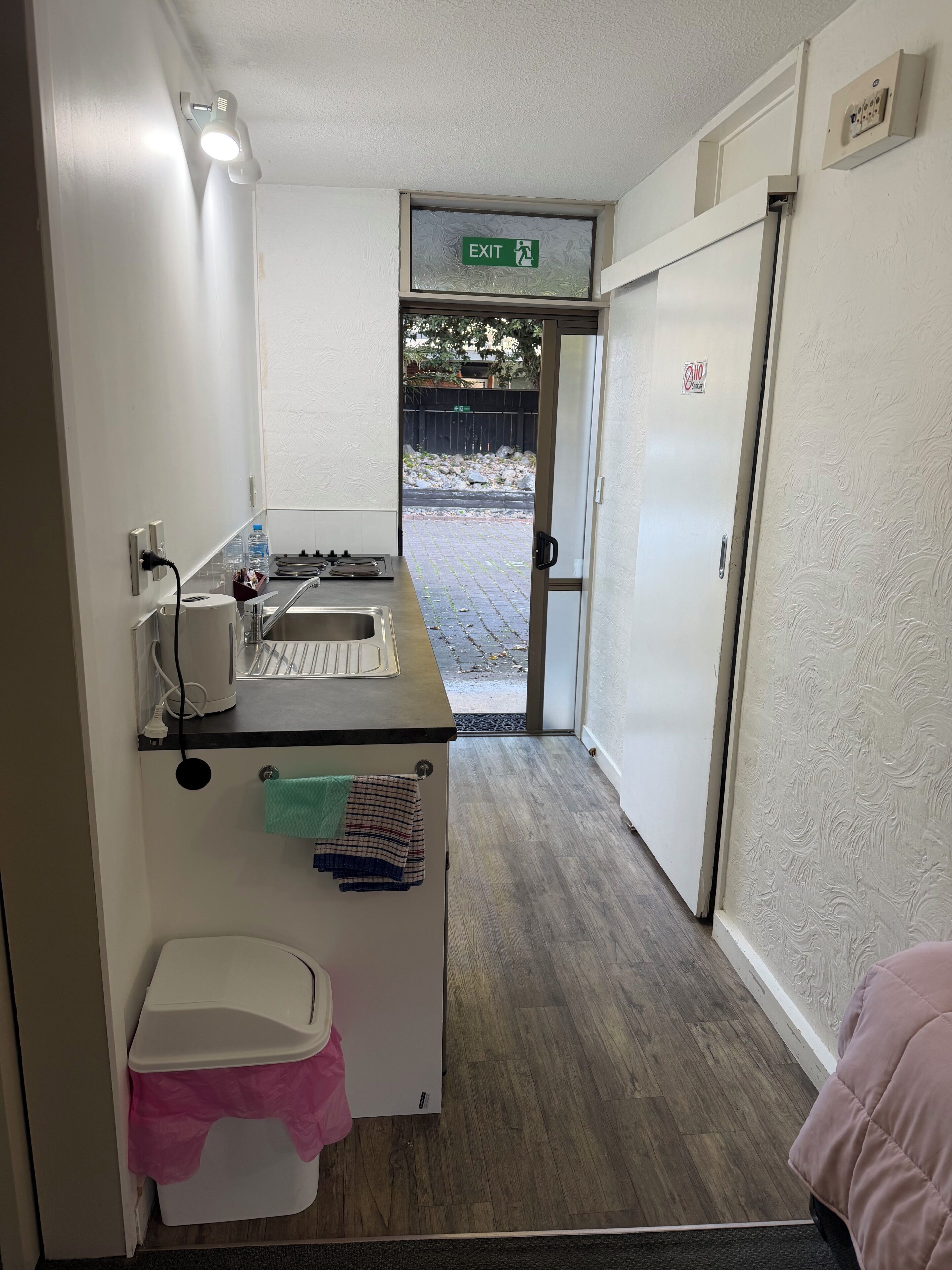 A narrow hallway with a kitchenette, a sink, a towel rack, a trash bin, and an exit sign.