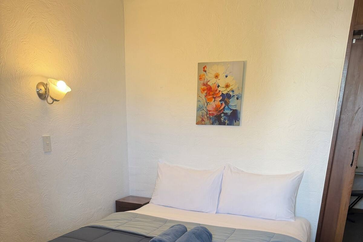 A neatly made bed with a blue blanket, a pair of rolled towels, and a painting of flowers on the wall.