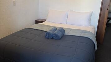 Een netjes opgemaakt bed met een blauwe deken, een paar opgerolde handdoeken en een bloemen schilderij aan de muur.