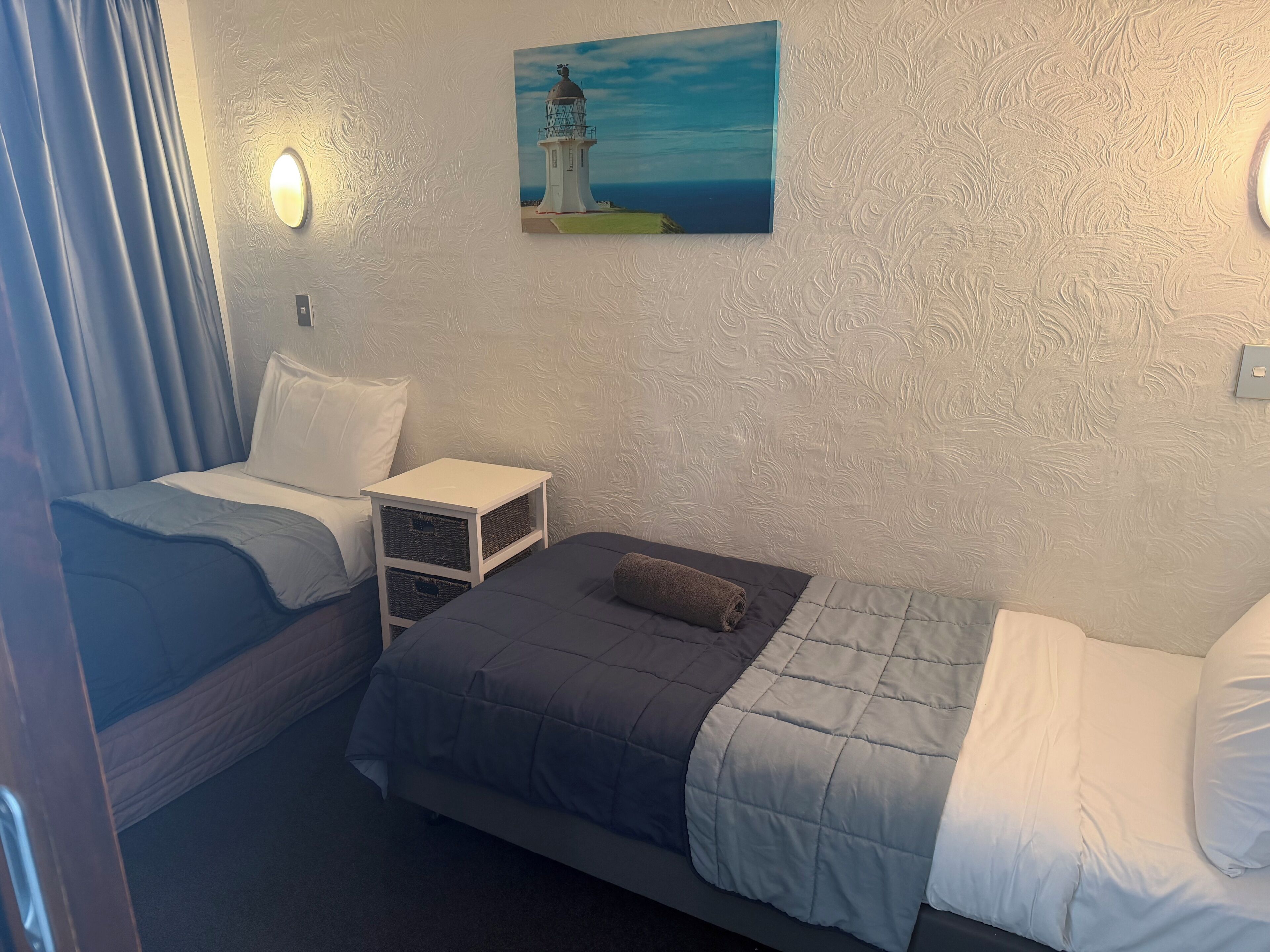 Un dormitorio con dos camas, una mesita de noche, un cuadro de un faro y una ventana con cortinas.