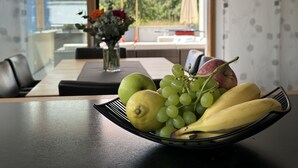 Una mesa de comedor con un frutero que contiene plátanos, uvas y manzanas. Hay un jarrón con flores y una vista de montañas a través de la ventana.