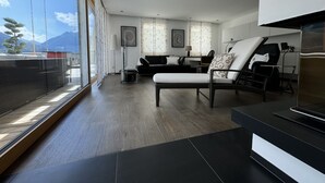 Una sala de estar moderna con un sofá, un sillón y un mueble para la televisión. Se ve una ventana grande con vistas a montañas.