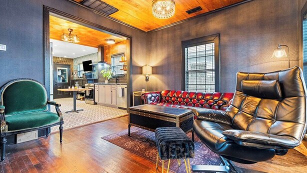 A spacious living room with a leather sofa, a green armchair, and a black leather chair. A wooden coffee table and a small ottoman are in the center. The room has wooden flooring and a large window with blinds. - Hollywood Vibe Cottage | Game Rm | Near Rainy St. (Austin)