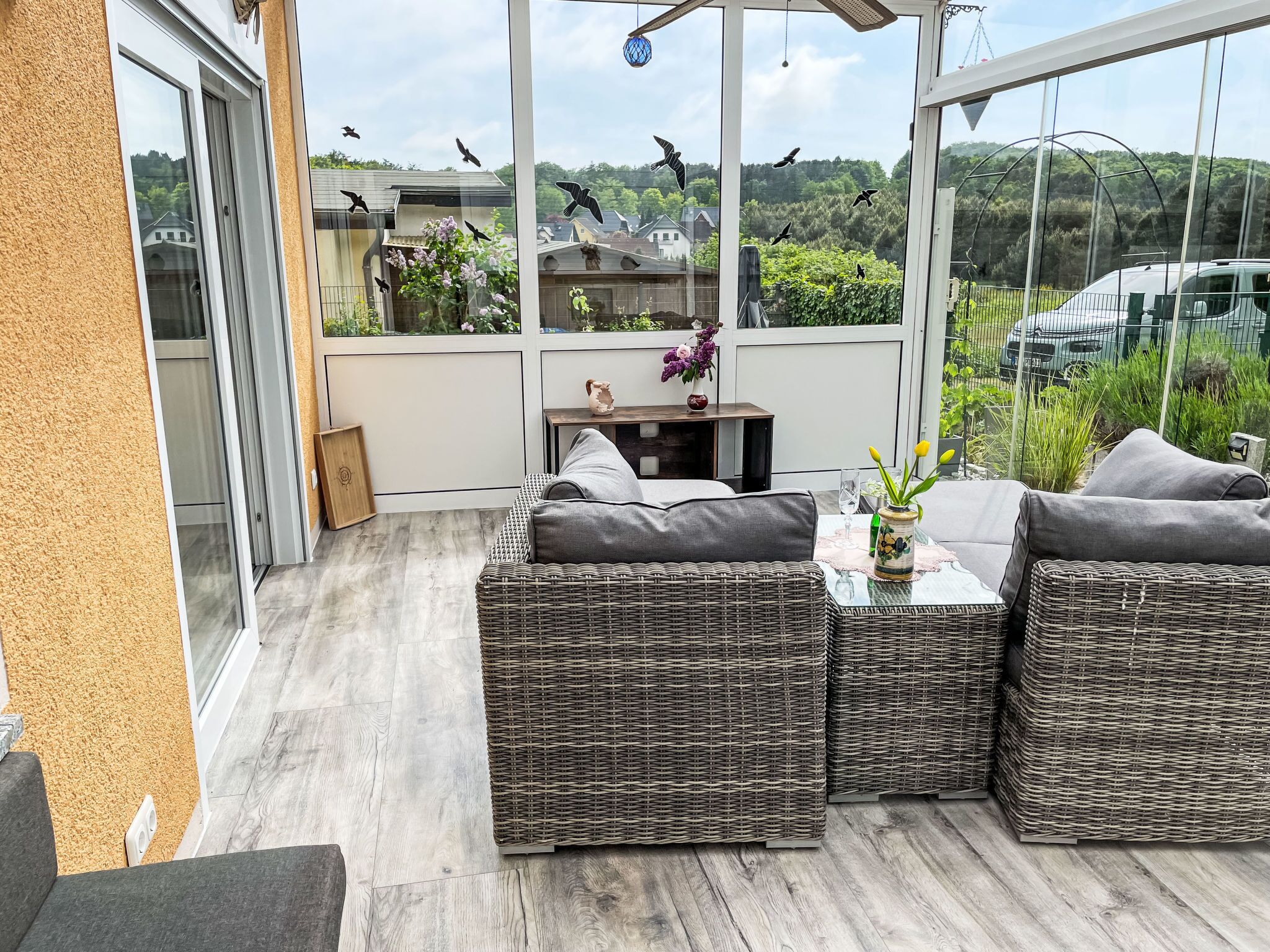 Un patio soleado con muebles de mimbre, una mesita con un florero y vistas a un paisaje verde y coches aparcados.