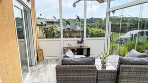 Une terrasse ensoleillée, meublée de meubles en osier, avec une petite table sur laquelle est posé un vase, et une vue sur un paysage verdoyant et des voitures garées.