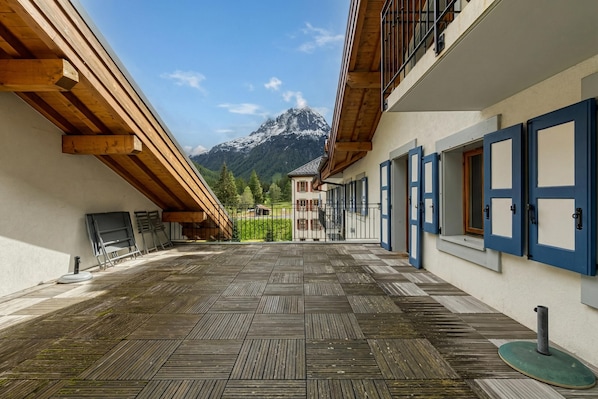 A covered outdoor area with a wooden floor, a view of a mountain, and a building with blue doors.