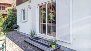 A house with a wooden exterior, a large window with white shutters, a barbecue grill, and a small wooden bench with a potted plant.