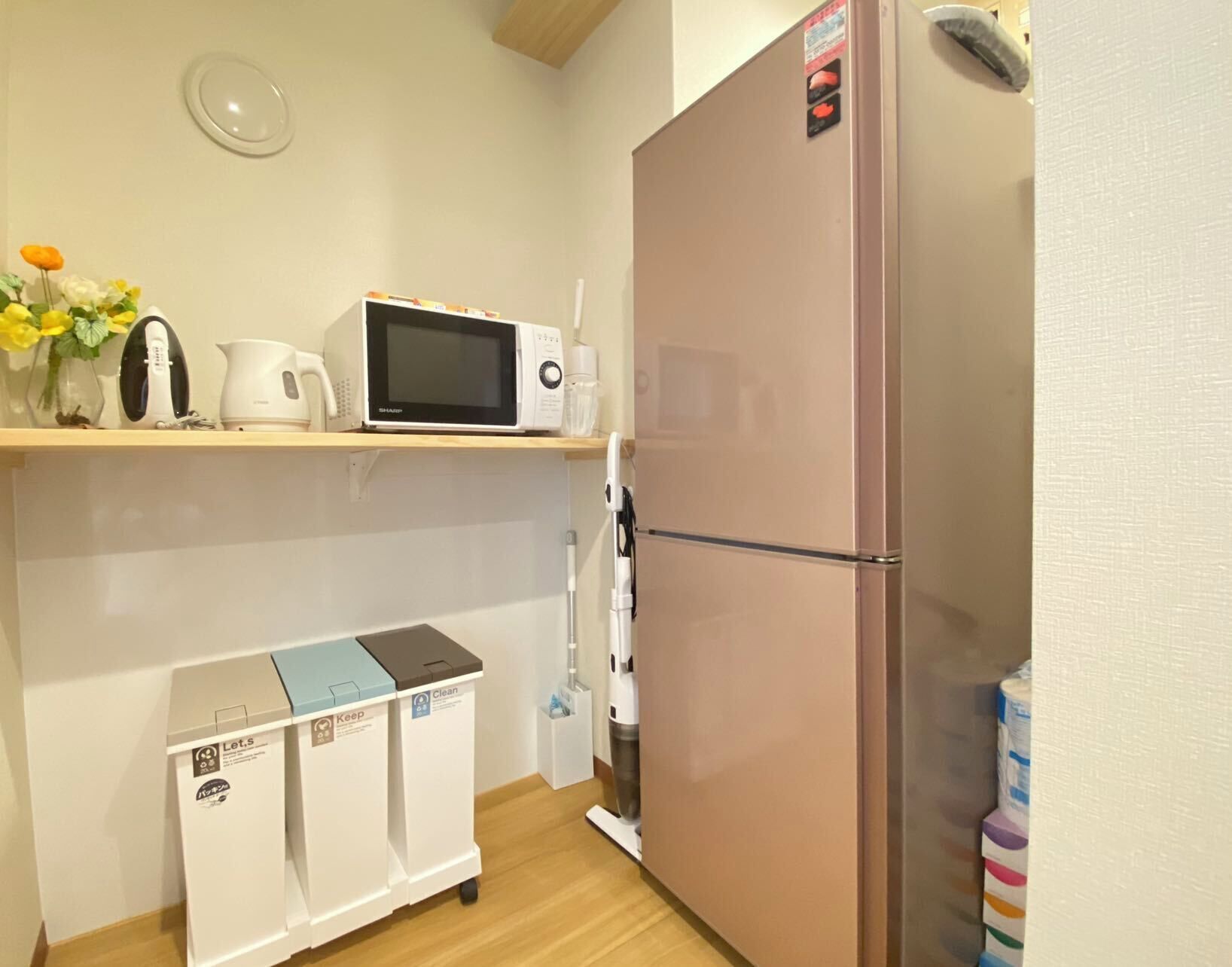 Cocina compacta con refrigerador, microondas, hervidor de agua y un estante pequeño con un hervidor y un jarrón de flores.