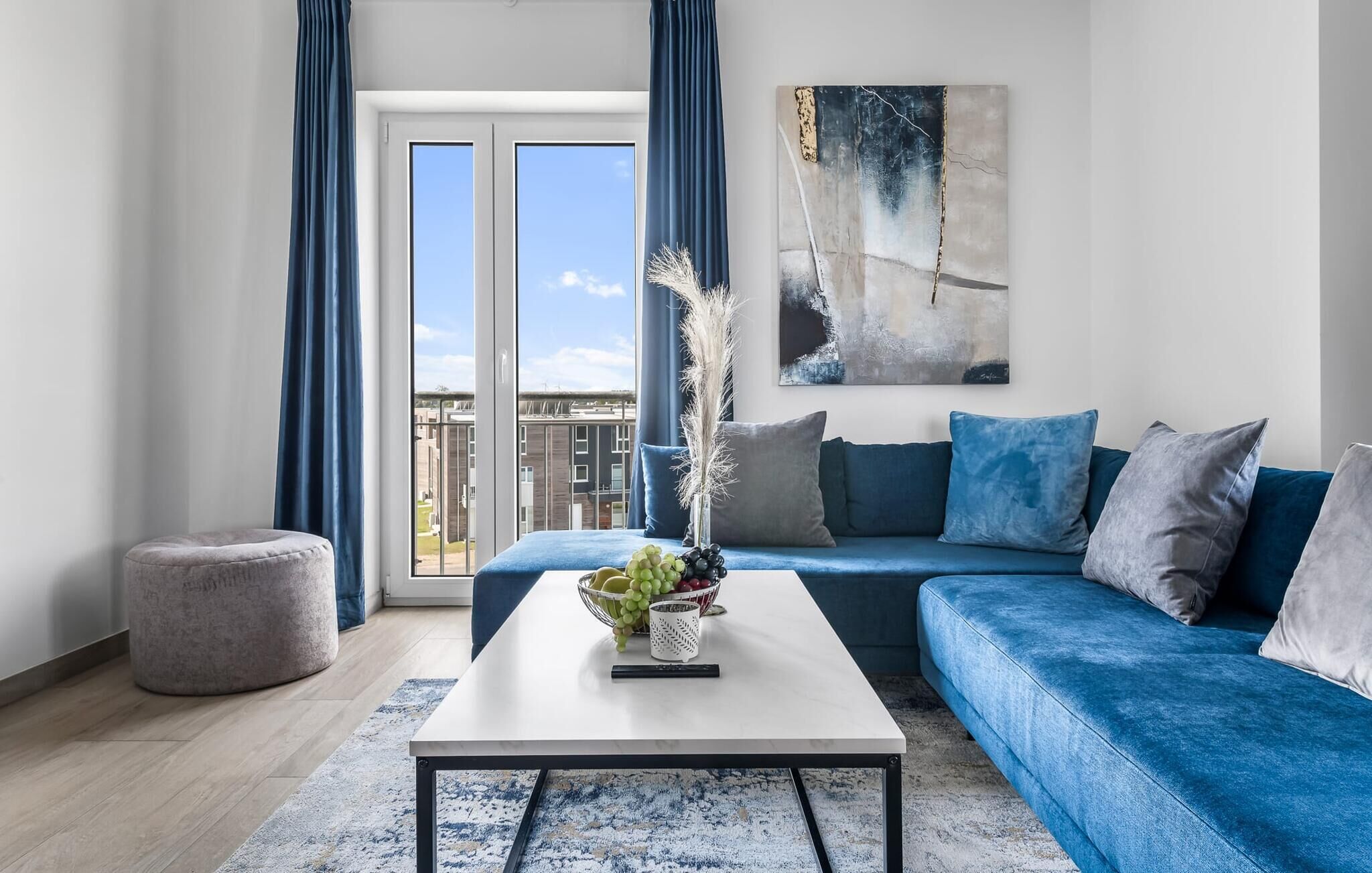 Un salon moderne avec un canapé bleu, une table basse blanche ornée de fruits et une vue sur la ville grâce à de grandes fenêtres.