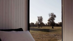A bedroom with a large window, a bed, and a view of a grassy field with trees. - Orana Cabin by Tiny Away (Cherry Tree Pool)