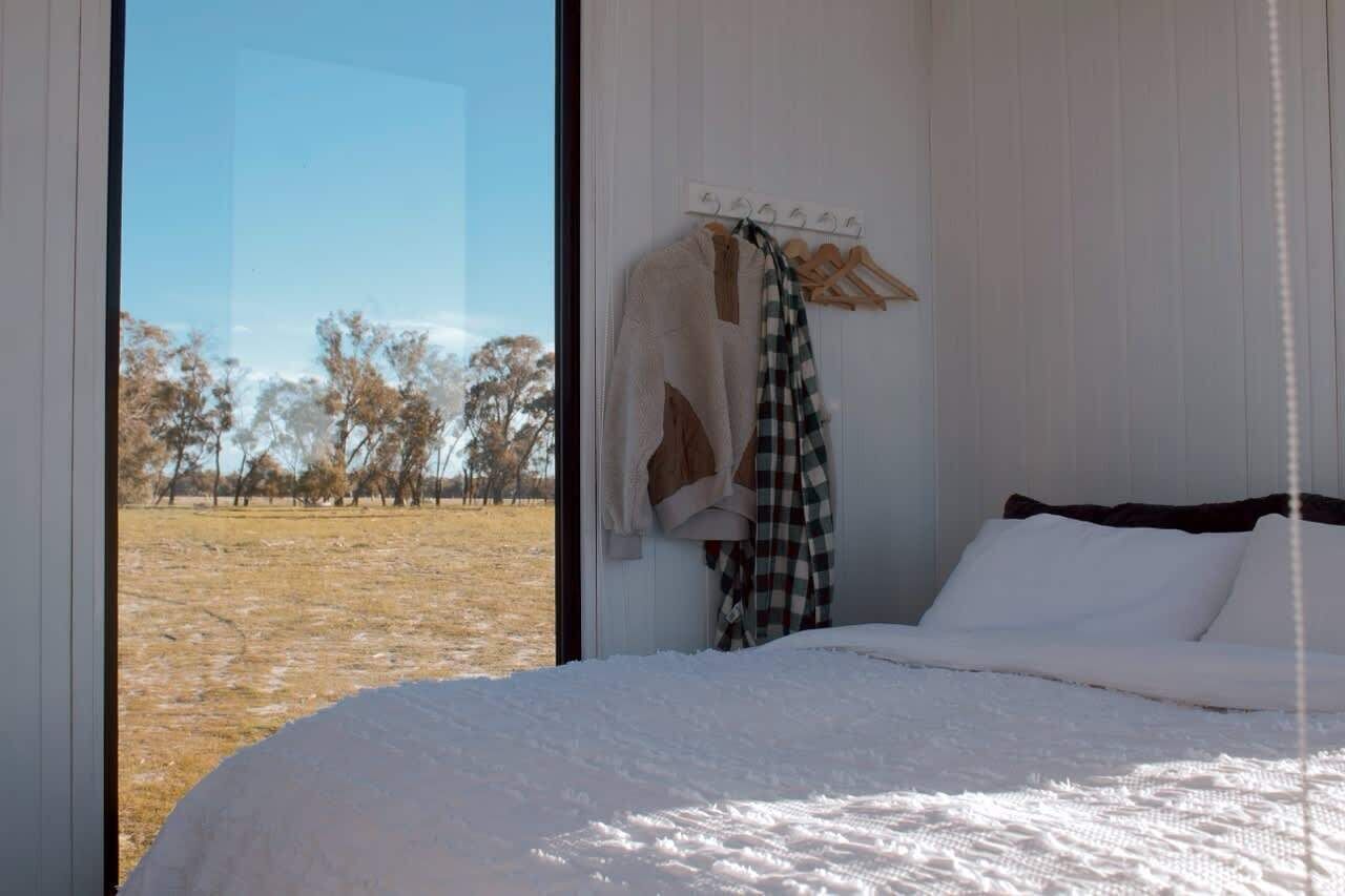 A bedroom with a large window, a bed, a hanging coat rack, and a view of a field.